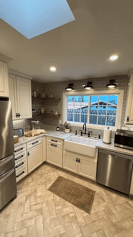 Modern kitchen with white cabinets, farmhouse sink, and herringbone tile flooring. Bright natural light.