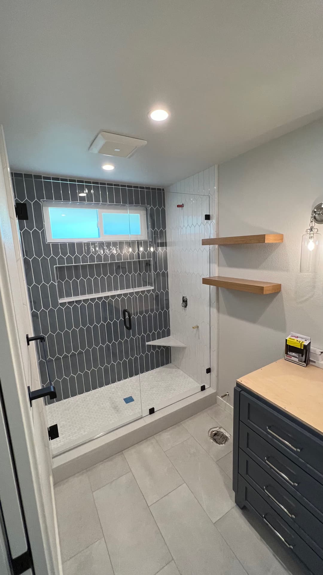 Modern bathroom with glass shower, gray tile accent wall, wood shelves, and stylish fixtures.